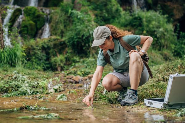 A person crouches down in front of a creek to collect a sample