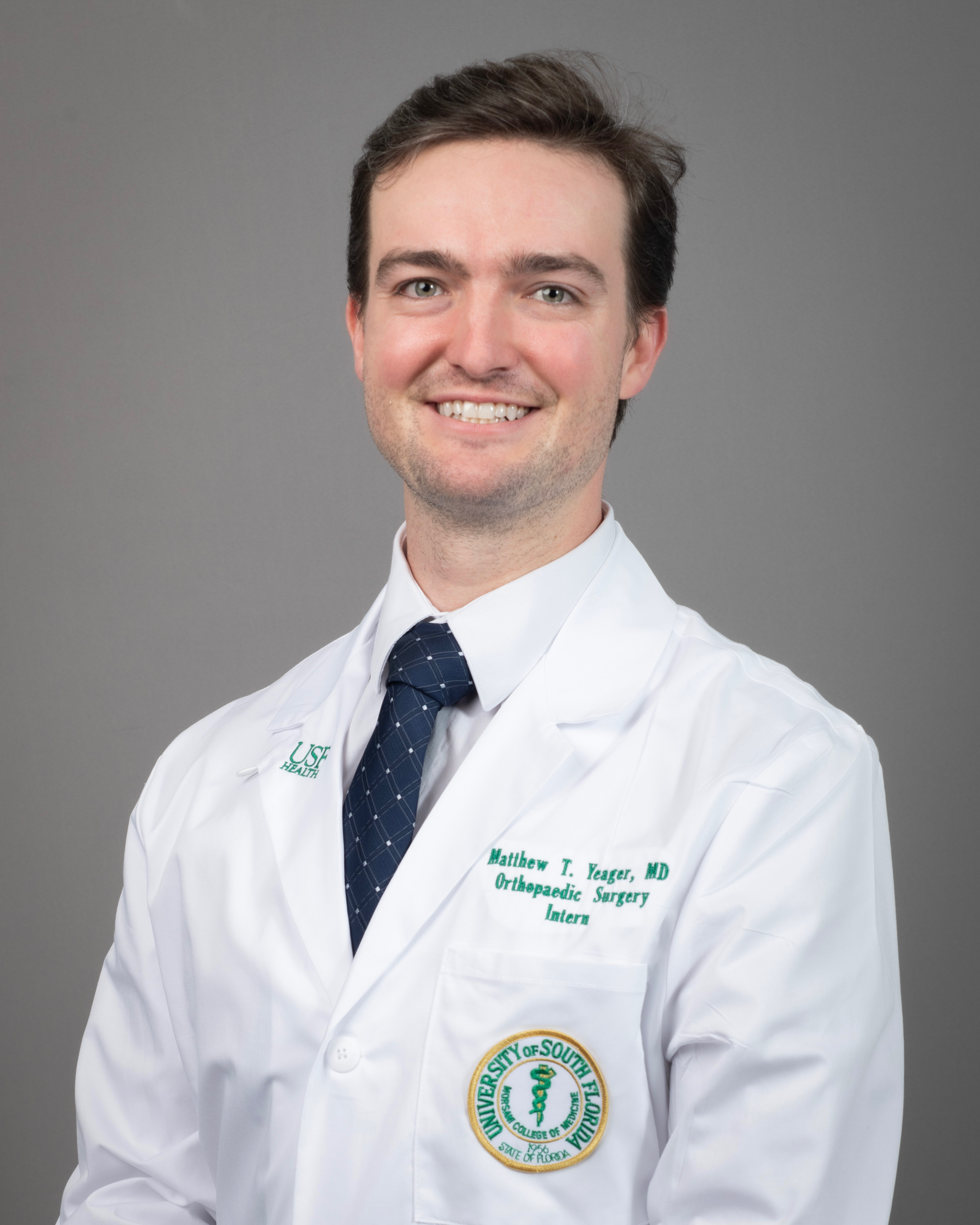 Matt smiling wearing a white dress shirt and navy blue tie wearing a USF Health branded medical white coat