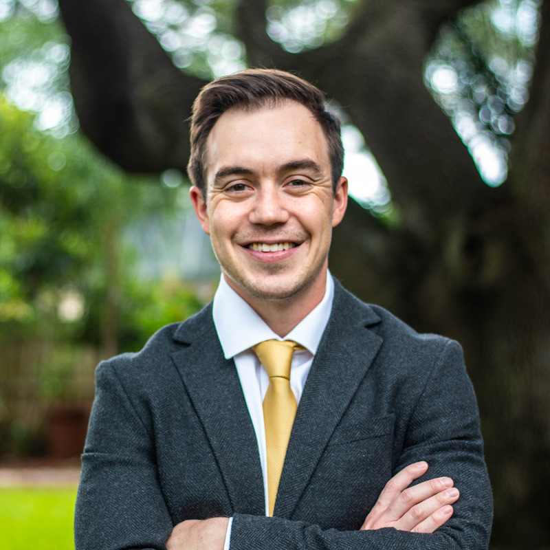 Cody Hawley smiling with his arms crossed wearing a dark grey suit with a gold tie, landscape background