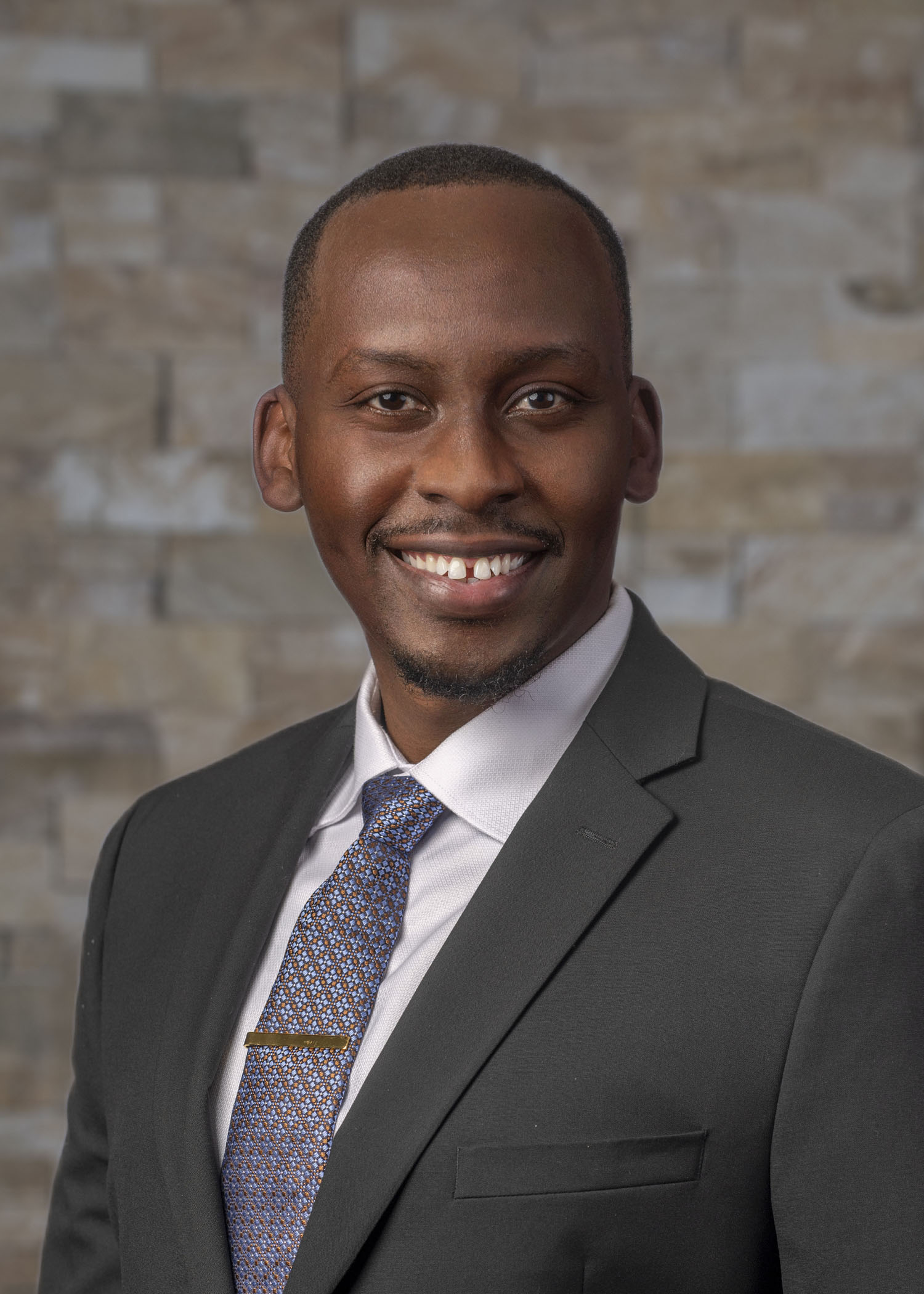 Headshot of Dr. Nsereko with closely cropped hair, wearing a dark suit, white shirt, and patterned blue tie against a stone wall background.
