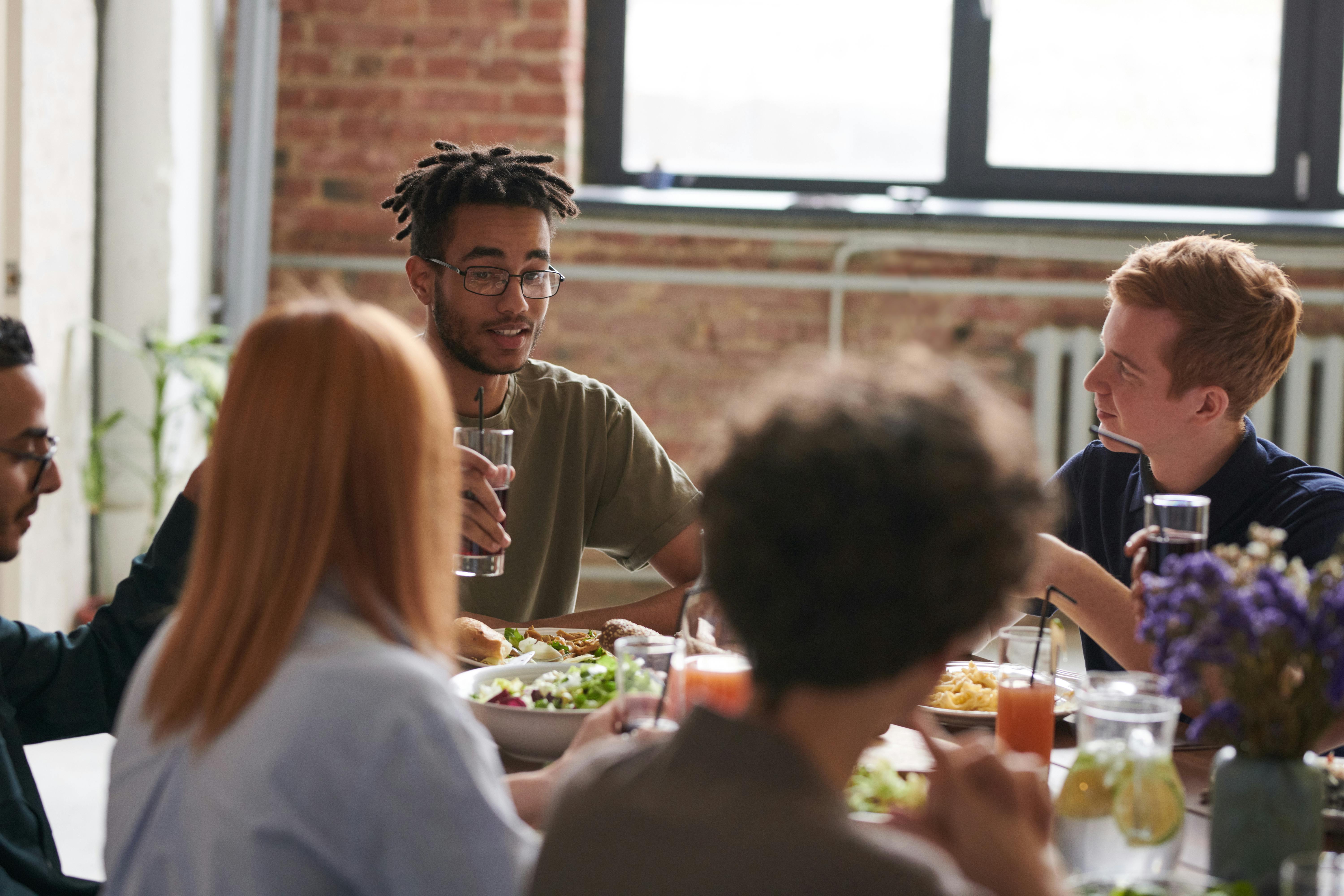 A group of young adults sitting around a table sharing a meal and drinks, engaged in conversation in a casual indoor setting with brick walls and natural light.