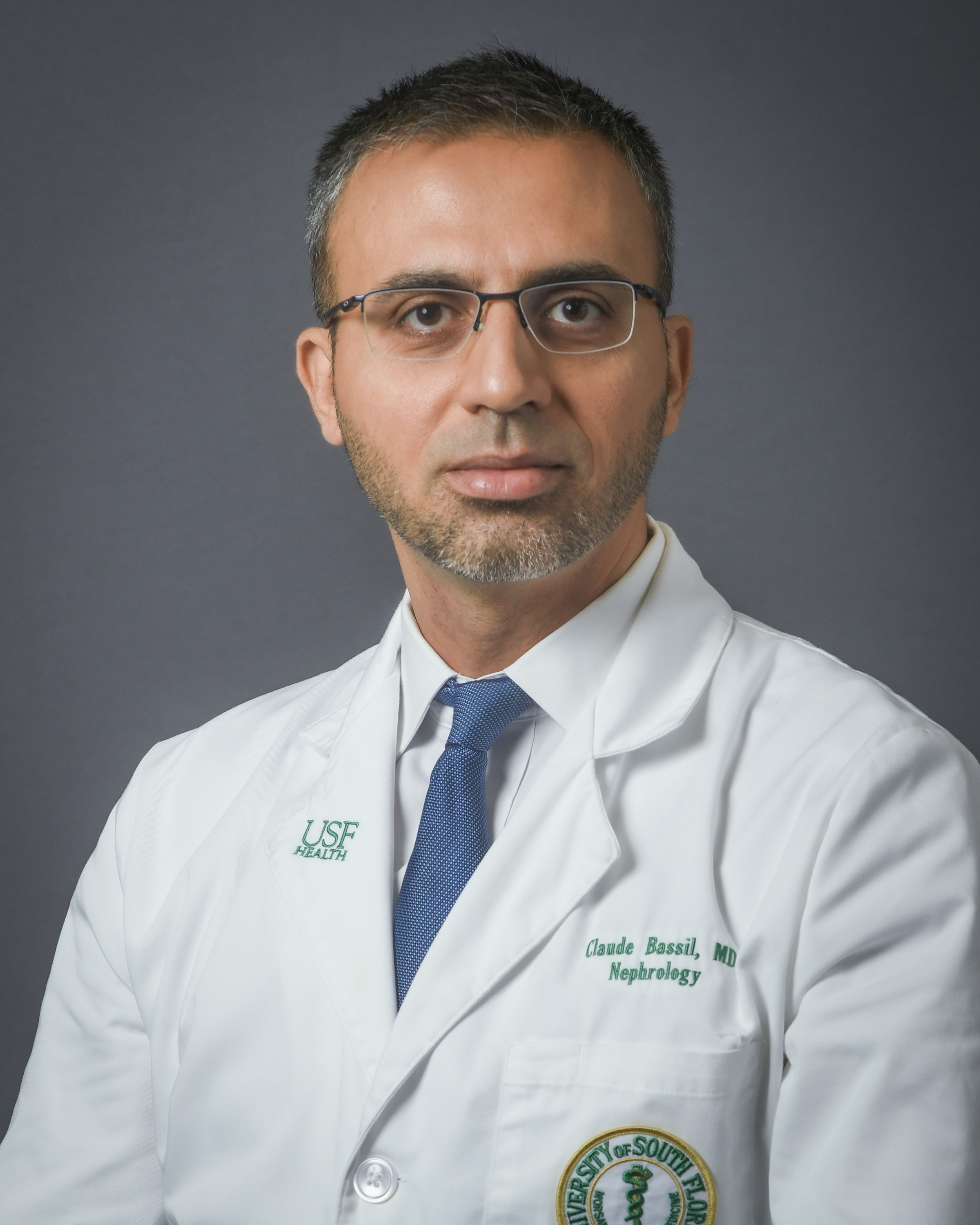 Dr. Claude Bassil posing for a headshot wearing a USF Health branded medical white coat with a dress shirt and blue tie