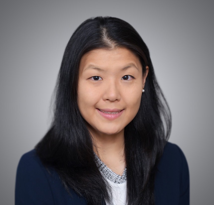 Headshot of Dr. Minha Kim with long dark hair, smiling slightly, wearing a dark blazer over a patterned top against a gray background.