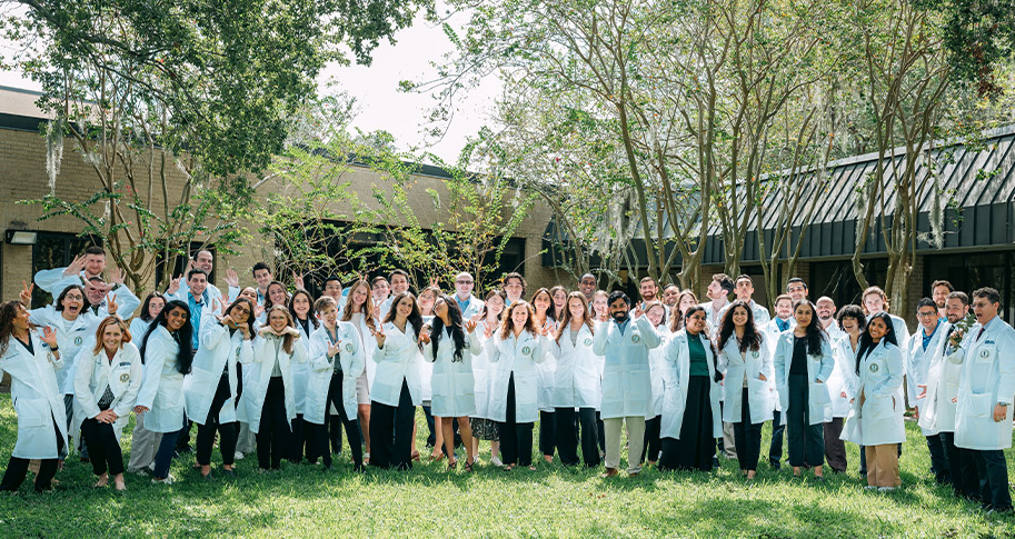 Large group shot of residents in their white coats, outside standing on grass with trees in the background.