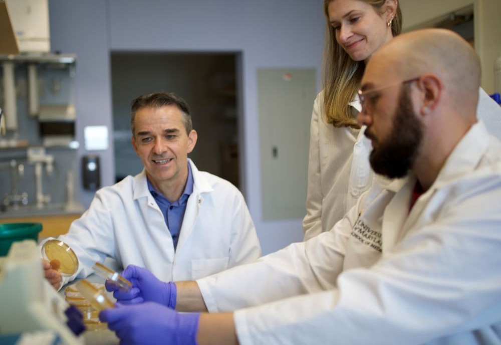 Dr. Ravel sitting at a desk with other medical professionals wearing white coats, looking at lab equipment