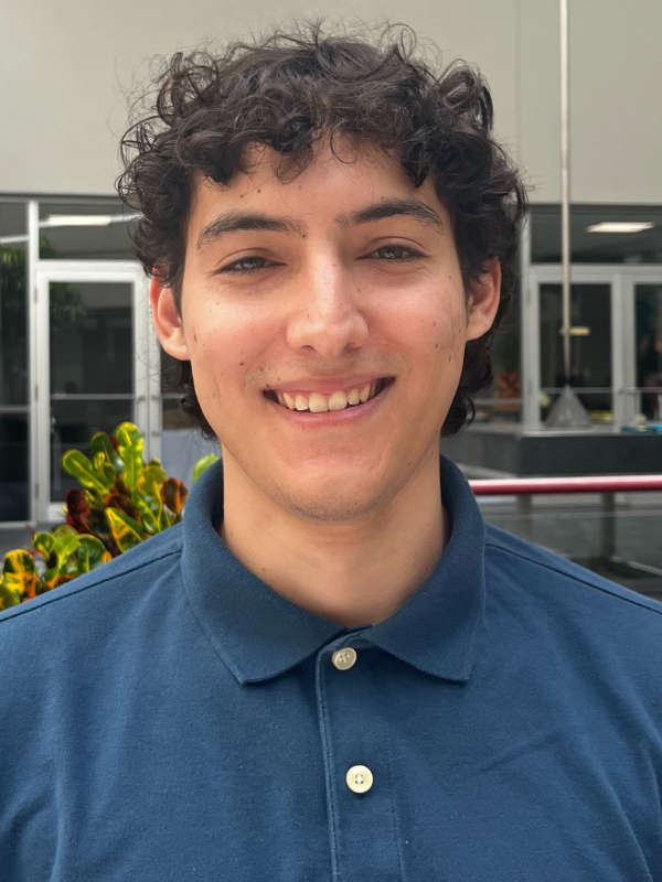 A headshot of Zachary Sanders in an outdoor setting. He is smiling and wearing a dark blue polo shirt.