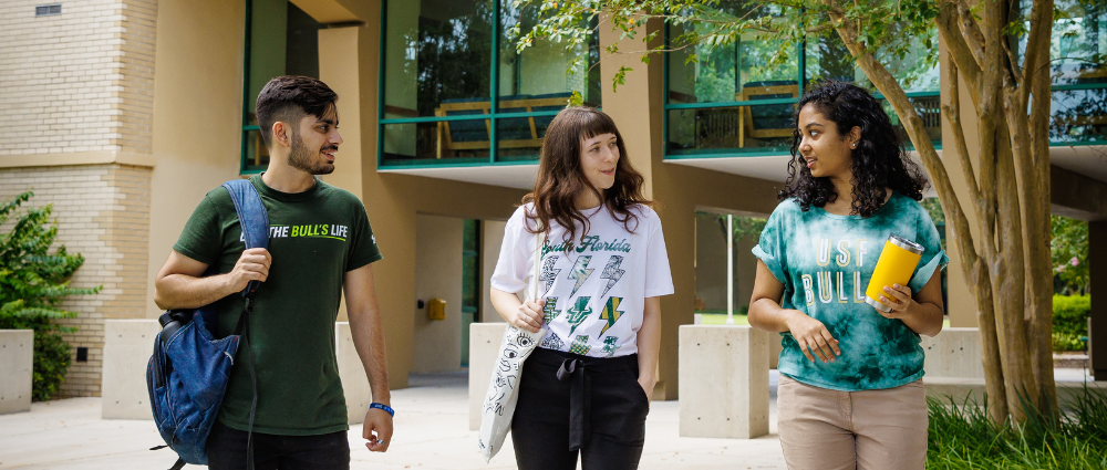 Three students walking outdoors on campus while chatting. They are wearing USF-branded t-shirts.