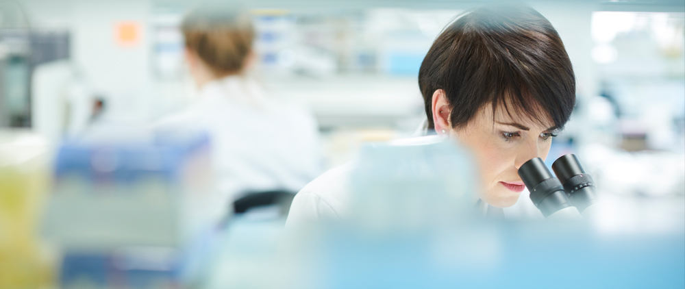 A female scientist in a lab coat is focused on observing a specimen under a microscope.