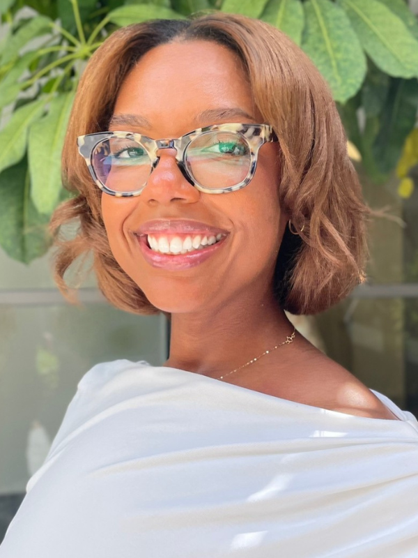 A headshot of Natalie Erasme smiling in front of greenery. She is wearing glasses and a white top.