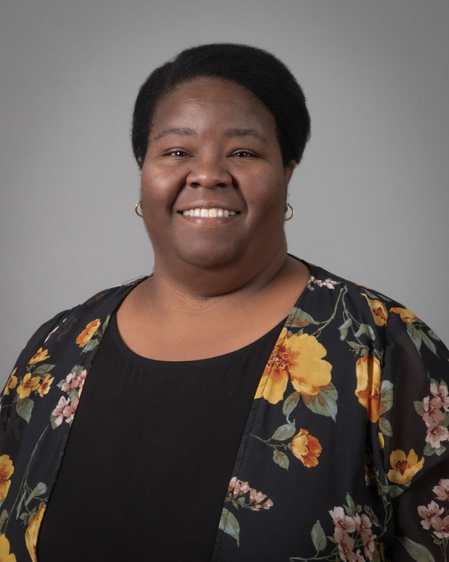 Monica Henderson smiling wearing a black blouse and floral shall in front of a grey backdrop