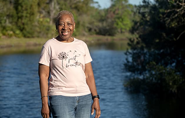 A woman stands by a river, gazing at the water with trees and a clear sky in the background.