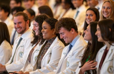 A large group of happy students wearing white coats sitting in an auditorium.