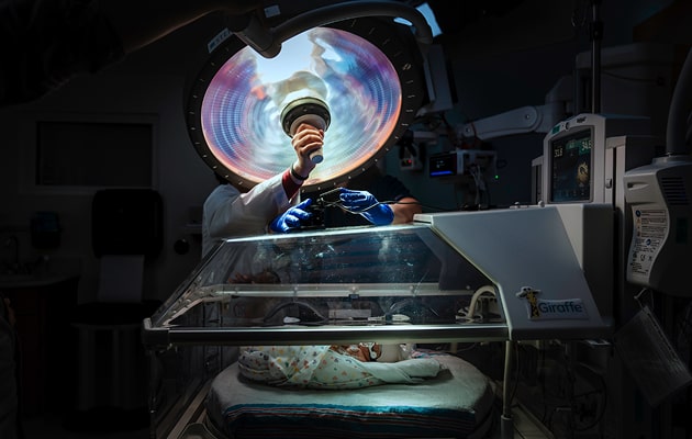 A medical professional conducts an examination on a baby in a hospital setting, with surgical tools and monitors visible.