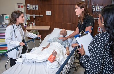 A nurse and two doctors converse over a high-fidelity manikin in a hospital bed, discussing care and treatment options.
