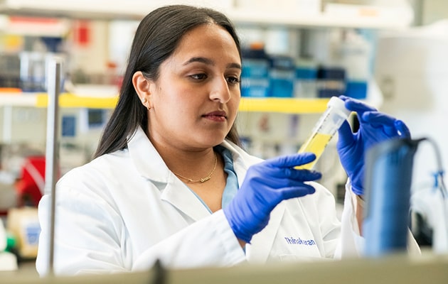 A closeup of a woman in a white lab coat, wearing blue gloves examines a test tube.